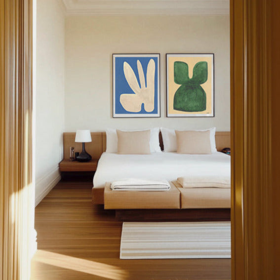Two framed abstract prints one blue with a white shape one yellow with a green shape above a bed with light wood frame